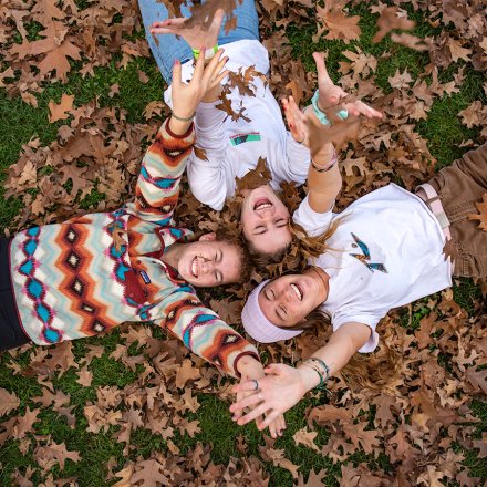 three students laughing while in a laying in a bed of fall leaves