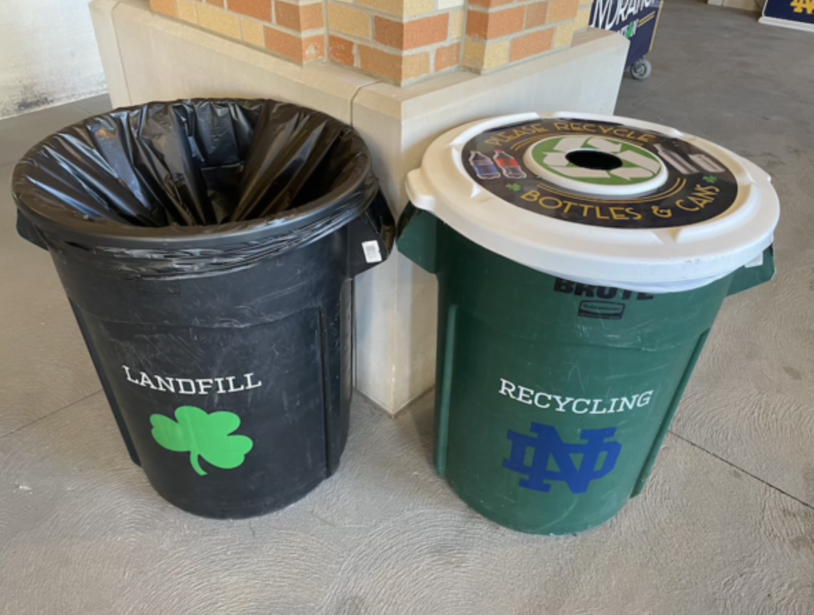 Stadium recycling and landfill barrels set up side-by-side: a green "RECYCLING" barrel for bottles, cups, and cans, and a black "LANDFILL" barrel for all other waste.