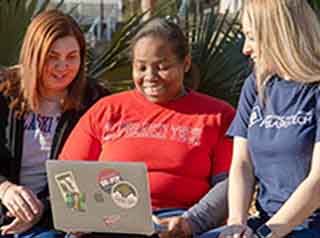 Three students sitting outside looking at their laptop screen.