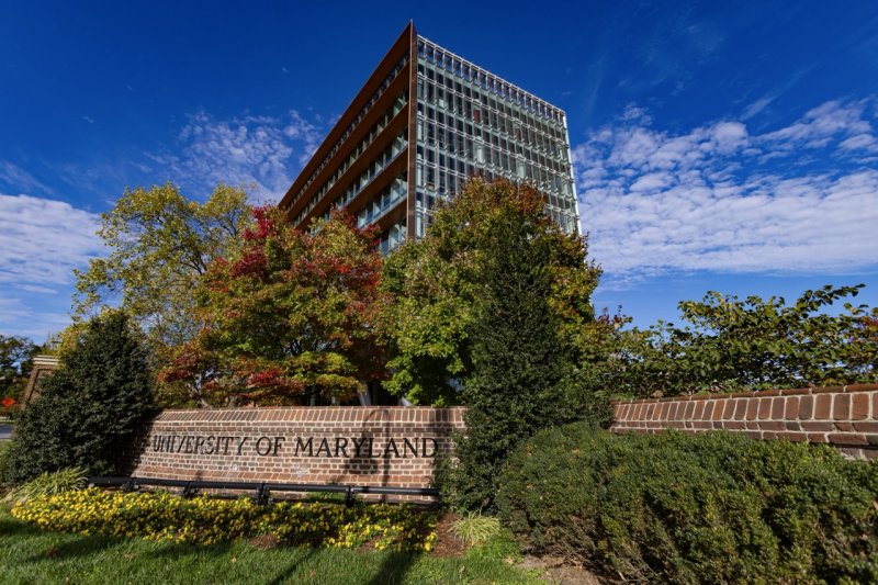 The sun shines on a University of Maryland sign at the entrance of the university near the Founder's Gate. Leaves on the trees behind the sign are a mix of green and red with a view of the Iribe Center in the background.