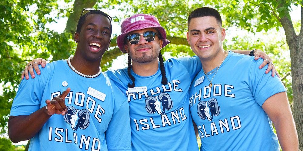 Three smiling students in matching &ldquo;Rhode Island&rdquo; T‑shirts stand outdoors with arms around each other