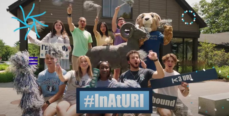 A group of cheerful URI students wearing matching blue shirts poses energetically outdoors under a blue sky, surrounded by greenery