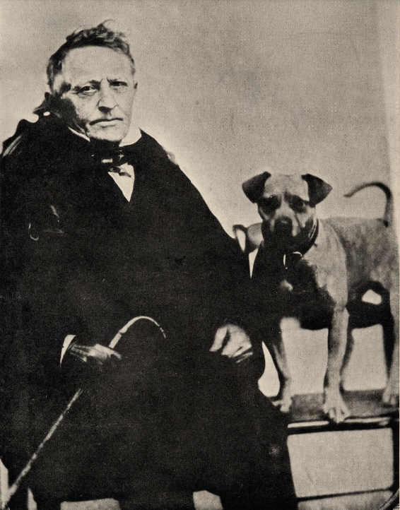 Three-quarter length picture of Matthew Vassar wearing glasses, black tie, white shirt, black suit and black gloves. He is seated, holding a curved-top cane in his right hand. His dog, Tip, is standing on a chair next to him.