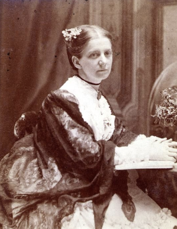 Three-quarter length picture of Eunice D. Sewall wearing flowers in her hair, black choker, broach, long, white ruffled dress and black lace shawl. She is seated, her arms resting on an open book.