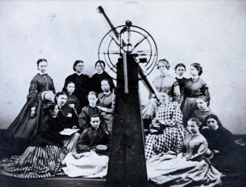 Professor Maria Mitchell and fifteen female students wearing long dresses, seated or standing behind a telescope. Some are holding hats or an open book.