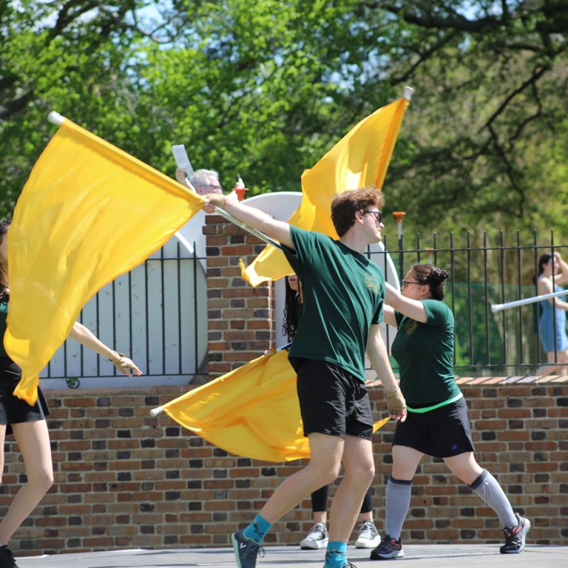People performing a color guard routine outdoors, waving large yellow flags in a coordinated formation near a brick wall and metal fence.