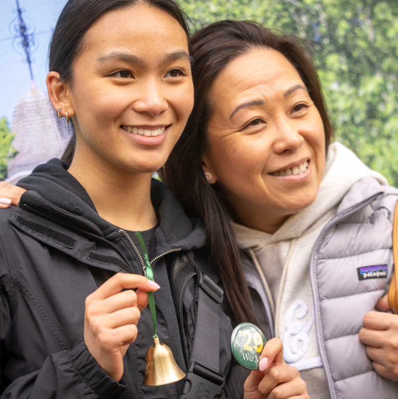 Two people smiling and posing together outdoors, one holding a small bell and a button.