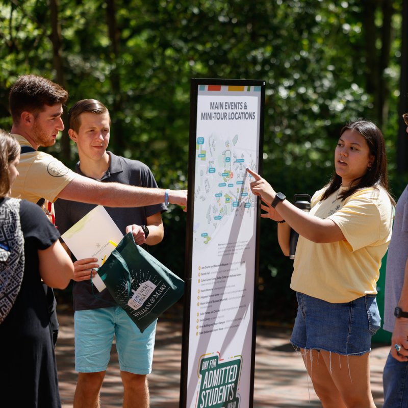 A group of students stands around a campus map sign labeled &ldquo;Main Events & Mini-Tour Locations,&rdquo; with one person pointing out locations while others look on, holding papers and a William & Mary tote bag.
