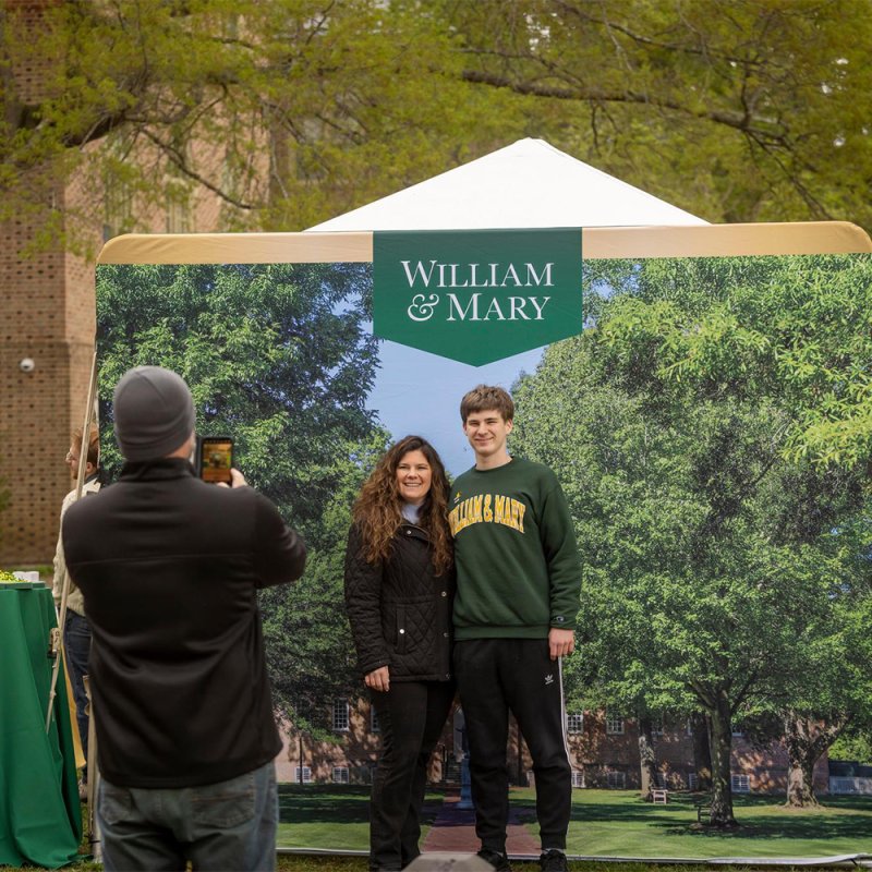 Two people pose for a photo while someone takes their picture in front of a campus backdrop.
