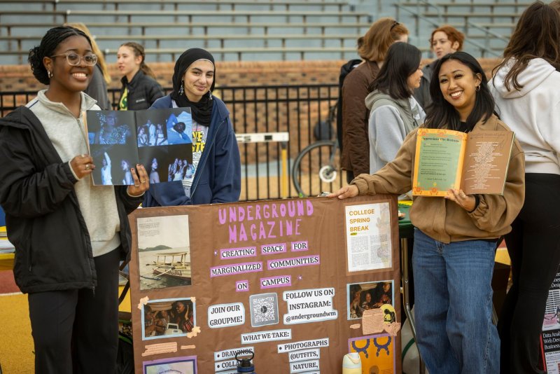 Students at a campus table hold open pages and stand behind a display board.