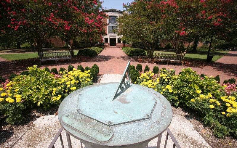 Bronze sundial outside Swem Library, centered in a flower bed along a brick path with benches and blooming trees.