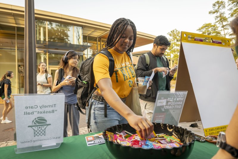 Students gather around an information table outside a campus building. A student wearing a yellow William & Mary T-shirt and backpack reaches into a bowl of candy while other students wait nearby. 